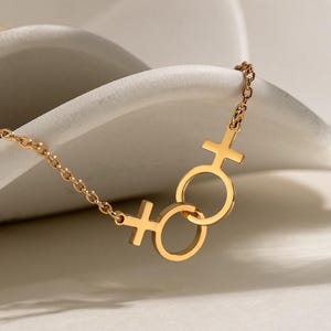 Może przedstawiać: Złoty naszyjnik z dwoma połączonymi symbolami żeńskimi. Łańcuszek jest delikatny, a zawieszka umieszczona jest na białym, zakrzywionym tle. Naszyjnik symbolizuje feminizm i wzmocnienie pozycji kobiet.