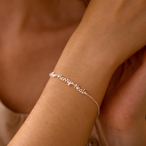 Peut inclure: Un bracelet argenté délicat avec les noms "Flo Henry Neals" en écriture cursive. Le bracelet est porté au poignet d'une personne, mettant en valeur le bijou personnalisé.