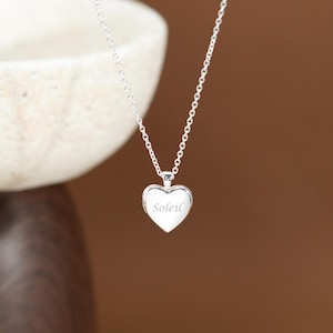 May include: A silver-toned heart-shaped locket pendant suspended from a delicate matching chain. The front of the heart features the word Soleil engraved in a cursive script. The metal has a polished finish that reflects light across its surface.