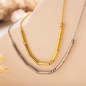 May include: Two minimalist necklaces, one gold and one silver. Each features a delicate chain with cylindrical beads. The necklaces are displayed on a neutral surface. The design is simple and elegant, suitable for everyday wear.