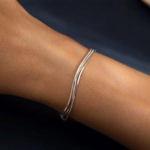 May include: A silver bracelet featuring a snake chain design. The bracelet comprises three strands of fine, textured metal, worn on a wrist against a dark blue backdrop. The jewellery is simple and elegant, suitable for everyday wear.