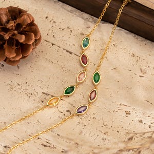 Könnte beinhalten: Zwei zarte goldene Halsketten mit farbigen Edelsteinakzenten. Jede Halskette hat eine Kette mit einer Reihe von oval geformten Edelsteinen in verschiedenen Farben, darunter Grün, Rot, Rosa und Lila. Die Edelsteine sind in Gold gefasst. Die Halsketten werden auf einer strukturierten Oberfläche präsentiert.