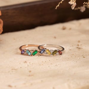 May include: Two rings with colourful gemstone accents. One ring is rose gold, the other is silver. Each ring features a row of rectangular gemstones in various colours, including purple, green, blue, yellow, and red. Small clear stones are interspersed.