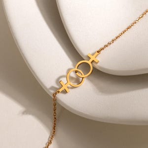 Może przedstawiać: Złota bransoletka z łańcuszkiem z przeplatającymi się symbolami Wenus. Bransoletka ma delikatny łańcuszek i polerowane wykończenie. Symbole Wenus są połączone małym pierścieniem. Bransoletka jest prezentowana na białej powierzchni.