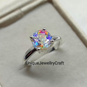 May include: A silver ring with a large, round, iridescent gemstone. The gemstone displays a spectrum of colors, including pink, yellow, and blue. The ring band is smooth and polished. The text "UniqueJewellryCraft" is visible.