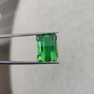 Könnte beinhalten: Ein leuchtend grüner Smaragd, geschliffen in Smaragdfassung, wird von einer Pinzette gehalten. Der Edelstein ist klar und hat einen brillanten Glanz.