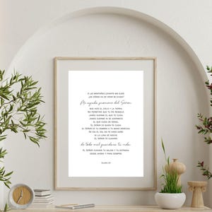 May include: A framed print with a light wood frame displays a poem in black script on a white background. The text includes the phrase "Mi ayuda proviene del Señor." The print is displayed on a wooden shelf with decorative plants.