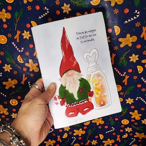 Könnte beinhalten: Eine weiße Karte mit einer Illustration eines Gnom mit roter Mütze und grünem Mantel, der eine Zuckerstange hält. Die Karte enthält den Text "Trova la magin in tutto ciò che ti circonda" und ein Laternen-Design. Der Hintergrund ist ein festliches Muster.