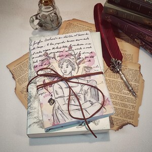 Op de afbeelding: Een gesloten dagboek met een bruine leren omslag en een lint eromheen gebonden. De omslag heeft een aquarelillustratie van een vrouw met een boek in haar hand. Het dagboek ligt op een tafel met een pen en een stapel boeken.