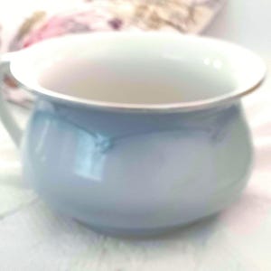 May include: A small, light blue and white ceramic chamber pot with a handle. The pot has a rounded body and a wide, flat rim. The interior is white, and the exterior is a soft, muted blue. The handle is curved and attached to the side of the pot.