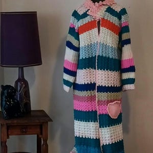 Könnte beinhalten: Ein langer, handgefertigter Häkelmantel in verschiedenen Farben, darunter Rosa, Blau, Orange und Weiß. Der Mantel hat eine herzförmige rosa Tasche und eine dekorative rosa Bordüre um den Kragen und die Kanten. Der Mantel wird auf einer Schaufensterpuppe präsentiert.
