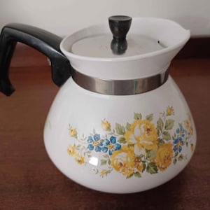 Könnte beinhalten: Weiße Keramik-Teekanne mit schwarzem Griff und Deckel. Die Teekanne ist mit gelben Rosen und blauen Blumen verziert. Ein silbernes Band umschließt die Mitte der Teekanne.