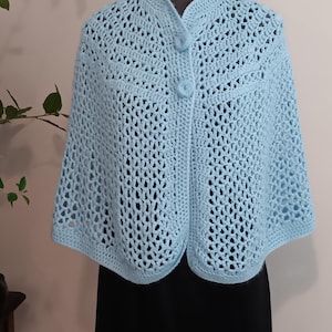 May include: A pale blue, crocheted cape with a button fastening. The open weave design creates a delicate, airy look. The cape is displayed on a mannequin, showcasing its shape and drape. The garment is a soft, pastel blue.
