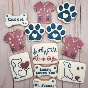 May include: A set of 10 decorated sugar cookies featuring a dog bone, paw prints, a dog silhouette, and a veterinarian's uniform. The cookies are decorated with blue, white, and pink icing. The text on the cookies reads "Cassie", "Logan Loves You", "Dr. Pounds", and "Thank You".