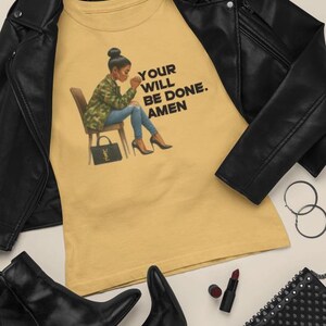 Op de afbeelding: Mosterdgeel T-shirt met de tekst "YOUR WILL BE DONE. AMEN" en een illustratie van een vrouw in een camouflagejas, jeans en hakken. Een zwarte leren jas, laarzen, zonnebril en een tas met studs staan ook op de foto.