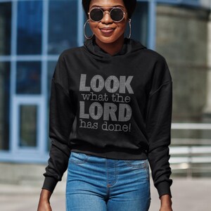 Puede incluir: Una sudadera con capucha negra con el texto "LOOK what the LORD has done!" en pedrería blanca.