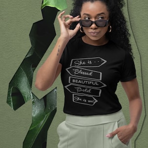 Może przedstawiać: Czarny t-shirt z napisem "She is Blessed, Beautiful, Bold, She is me" w białym wzorze strzałki. Osoba nosi okulary przeciwsłoneczne i jasnozielone spodnie. Tło to zielony abstrakcyjny wzór.