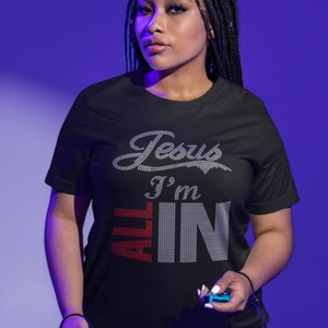 以下が含まれることがあります： 黒いTシャツに「Jesus I'm All In」という文字が銀色のグリッターで書かれており、「All」という単語は赤色のグリッターで書かれています。