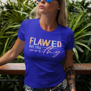 Op de afbeelding: Een koninklijk blauw T-shirt met de tekst "Flawed but still worthy" in regenboogkleuren. De tekst is in een speels lettertype en heeft een roze swirl eronder.