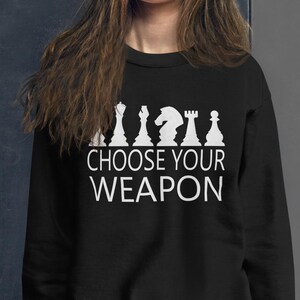 May include: Black sweatshirt with white chess piece silhouettes and the text "CHOOSE YOUR WEAPON". The sweatshirt is worn by a person with long brown hair and blue jeans. The background is a dark gray wall.