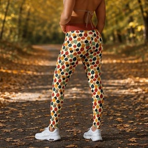 May include: Full-length leggings featuring a repeating pattern of small, teardrop-shaped leaves in shades of red, orange, yellow, green, and dark blue against a cream background, paired with a solid rust-coloured waistband and white athletic trainers.