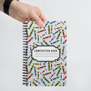 Op de afbeelding: Een blauw schrift met een spiraalbinding en een kleurrijk kleurpotloodpatroon. De omslag heeft een wit label met de tekst "COMPOSITION BOOK" in zwart.