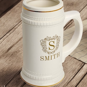 Könnte beinhalten: Ein weißer Bierkrug aus Keramik mit goldener Verzierung und einem goldenen Wappen mit dem Buchstaben "S" und dem Namen "SMITH".