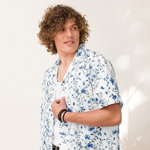 Puede incluir: Una camisa blanca de manga corta con un estampado floral azul. La camisa tiene cuello y cierre de botones en la parte delantera.