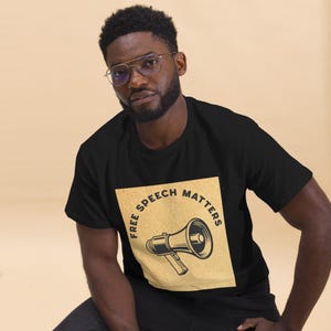 Puede incluir: Camiseta negra con un diseño gráfico cuadrado de color beige que presenta un megáfono y el texto "FREE SPEECH MATTERS" en un diseño circular. La camiseta está hecha de un material suave y tiene un cuello redondo clásico.