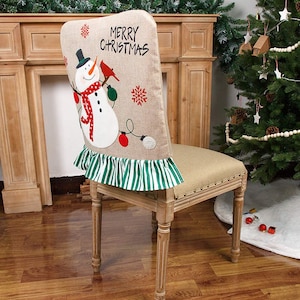 May include: A beige chair back cover with a snowman and robin applique. The cover has a green and white striped ruffle and the text "Merry Christmas".