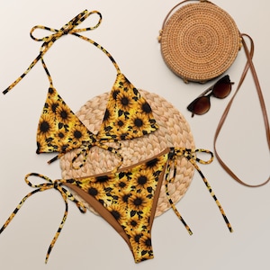 May include: A two-piece bikini set with a sunflower pattern. The top is a triangle style with tie straps, and the bottoms are a classic cut with tie sides. A round woven bag, sunglasses, and a woven placemat are also in the image.