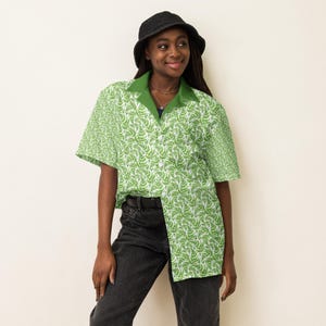 May include: A short-sleeved button-up shirt with a green fern pattern on a white background. The shirt has a green collar and is worn with black jeans and a black bucket hat. The shirt is made of a lightweight fabric.