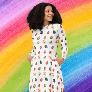 Puede incluir: Un vestido blanco con un estampado de huevos de Pascua coloridos. El vestido tiene mangas largas y un escote redondo. La modelo lleva el vestido y está de pie frente a un fondo de arcoíris.