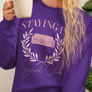 Puede incluir: Una sudadera morada con el texto "Staying In Social Club" en rosa claro. El diseño incluye una ilustración de sofá rosa dentro de una corona de hojas. La sudadera es de cuello redondo.