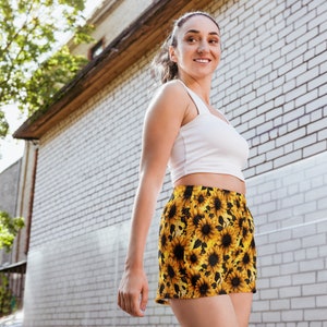 Puede incluir: Una mujer con un top blanco sin mangas y pantalones cortos de estampado de girasol negro y amarillo. Está de pie frente a una pared de ladrillo blanco.