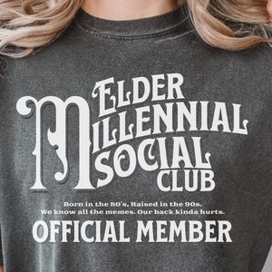 以下が含まれることがあります： 「Elder Millennial Social Club」という白い文字が書かれた濃いグレーのTシャツ。文字はビンテージスタイルのフォント。文字の下には、さらに小さな白い文字で「Born in the 80's, Raised in the 90s. We know all the memes. Our back kinda hurts. OFFICIAL MEMBER」と書かれている。