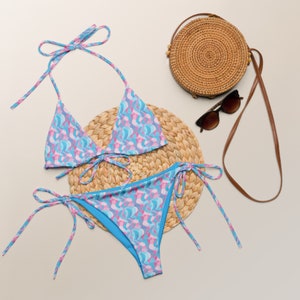 May include: A blue and pink patterned bikini with a matching top and bottom. The bikini is on a woven straw mat with a brown wicker bag and sunglasses.