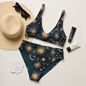 May include: A two-piece swimsuit with a celestial design featuring gold suns, moons, and stars on a navy blue background. The swimsuit is accompanied by a straw hat, sunglasses, sunscreen, and a seashell.