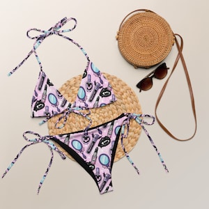 Puede incluir: Un bikini rosa y negro con estampado de labios, espejos y botellas de pintalabios. La parte superior del bikini es de estilo triangular con tirantes ajustables. La parte inferior del bikini es de estilo de corte alto con lazos en los lados.