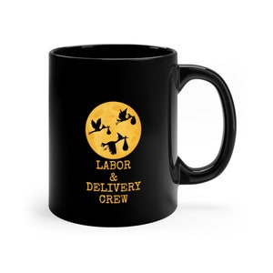 Può includere: Tazza in ceramica nera con una luna gialla e tre cicogne nere che portano bambini. Il testo "LABOR & DELIVERY CREW" è stampato sotto la luna.