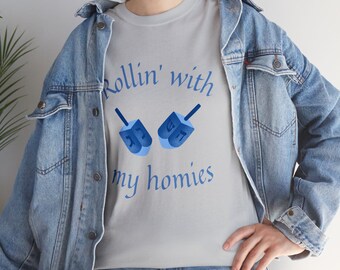 Könnte beinhalten: Hellgraues T-Shirt mit dem blauen Schriftzug "Rollin' with my homies" und zwei blauen Dreidels. Das Shirt wird mit einer Jeansjacke getragen.