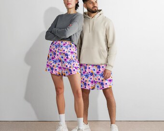 May include: A woman and a man stand side-by-side, both wearing matching floral print shorts. The woman wears a gray long-sleeve shirt, while the man wears a beige hoodie. The shorts feature a pink background with blue, purple, and white flower designs.