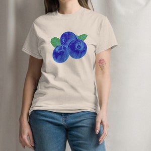 May include: A light beige t-shirt with a graphic of three blue blueberries and green leaves. The blueberries are drawn in a vintage style.