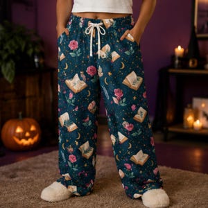 Op de afbeelding: Donkerblauwe loungebroek met een herhalend patroon van open boeken, roze rozen, gele maansikkels en kleine sterren. De broek heeft een wit trekkoord bij de elastische tailleband en twee zijzakken, wat zorgt voor een comfortabele, losse pasvorm.