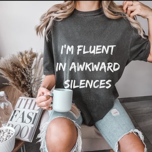 Puede incluir: Camiseta gris oscuro con el texto "I'M FLUENT IN AWKWARD SILENCES" en blanco. La persona sostiene una taza azul claro. La persona lleva vaqueros azules rotos. Se ve un libro titulado "THE FASHION BOOK".
