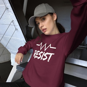 Può includere: Una felpa color borgogna con una grafica bianca di una linea di battito cardiaco e la parola "RESIST" in un carattere grassetto e stilizzato. La modella indossa un berretto da baseball grigio.