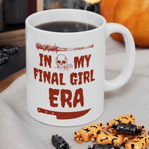 以下が含まれることがあります： 白いセラミック製のコーヒーカップに、赤い文字で「In My Final Girl Era」と書かれています。デザインには、口にバラを持った骸骨、有刺鉄線の野球バット、ナイフが含まれています。