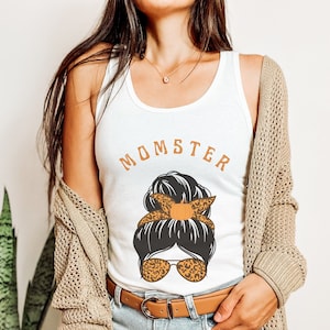 以下が含まれることがあります： オレンジ色の文字で「Momster」と書かれた白いタンクトップ。テキストの下には、乱れたお団子ヘア、サングラス、髪にカボチャをつけた女性の漫画イラストがあります。