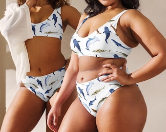 Pode incluir: Duas mulheres vestindo roupas de banho brancas e azuis com padrão de tubarão e peixe. Os trajes de banho são um top de biquíni e uma parte de baixo de cintura alta.