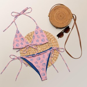 May include: A pink bikini with a blue seashell and starfish print, featuring tie-side bottoms and a triangle top with string ties. Included are a round woven straw bag with a long brown strap and dark-tinted sunglasses with brown frames.